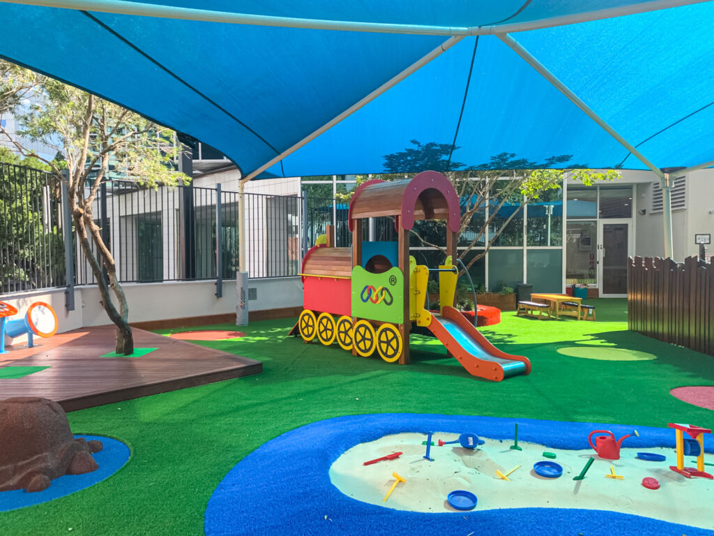 Sheltered outdoor playground at Montessori Academy’s Chatswood Childcare Centre, with a sand pit, mini-slide and turfed play areas.