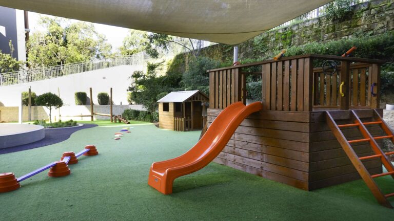 Outdoor playground at Montessori Academy’s Lane Cove West childcare, with a slide, playhouse and turfed play areas.