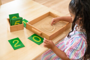 Young girl with Montessori Mathematics Material tracing Sandpaper Numbers in sand