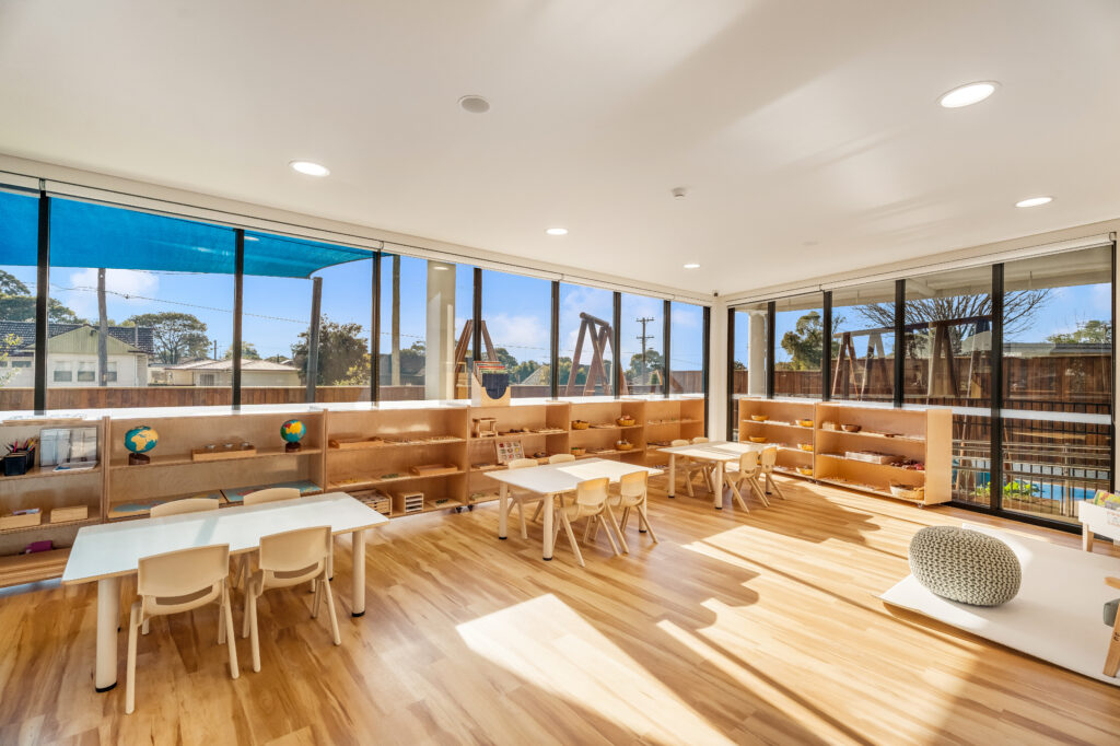 Classroom at Montessori Academy's St Marys childcare centre with three tables, each surrounded by four chairs, and shelves in the background.