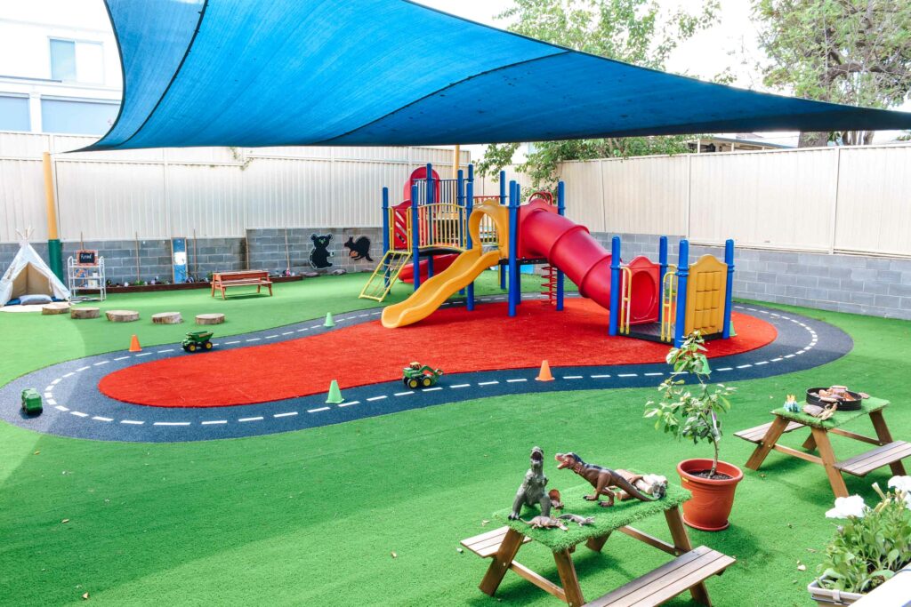 Sheltered outdoor playground at Montessori Academy’s Auburn childcare, with turfed play areas, slides and children’s tables.