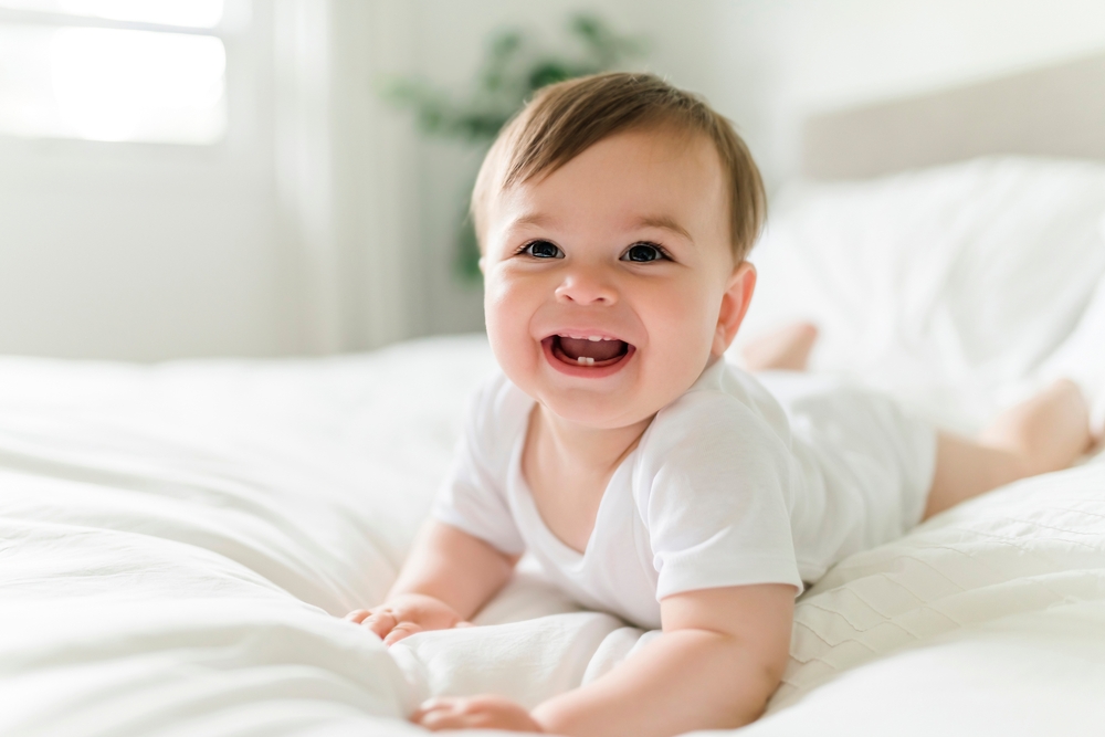 cute and smiling baby lying on white bedsheets in sunlit room