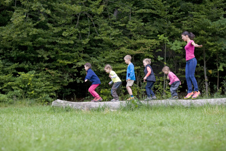 Kindergarten Teacher Playing With Kids On Log In A Wood