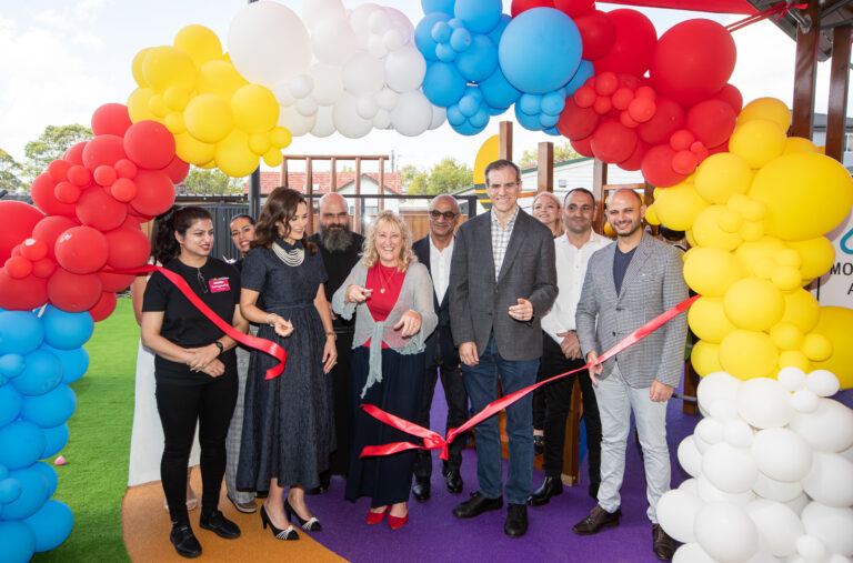 A group of people, including community leaders and educators, stand beneath a colourful balloon arch behind a ribbon, celebrating the grand opening of a new Montessori childcare centre in Panania, NSW.