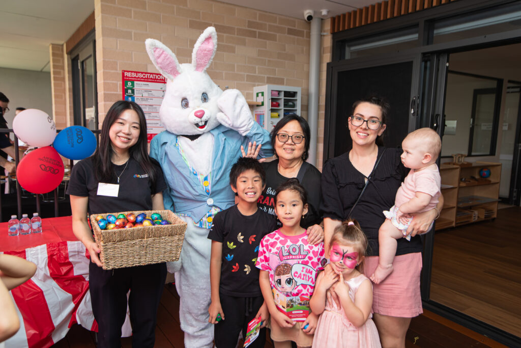 The Easter Bunny poses with young families and a Montessori Academy team member holding a basket of chocolate Easter eggs, celebrating the grand opening of the Panania childcare and preschool centre.