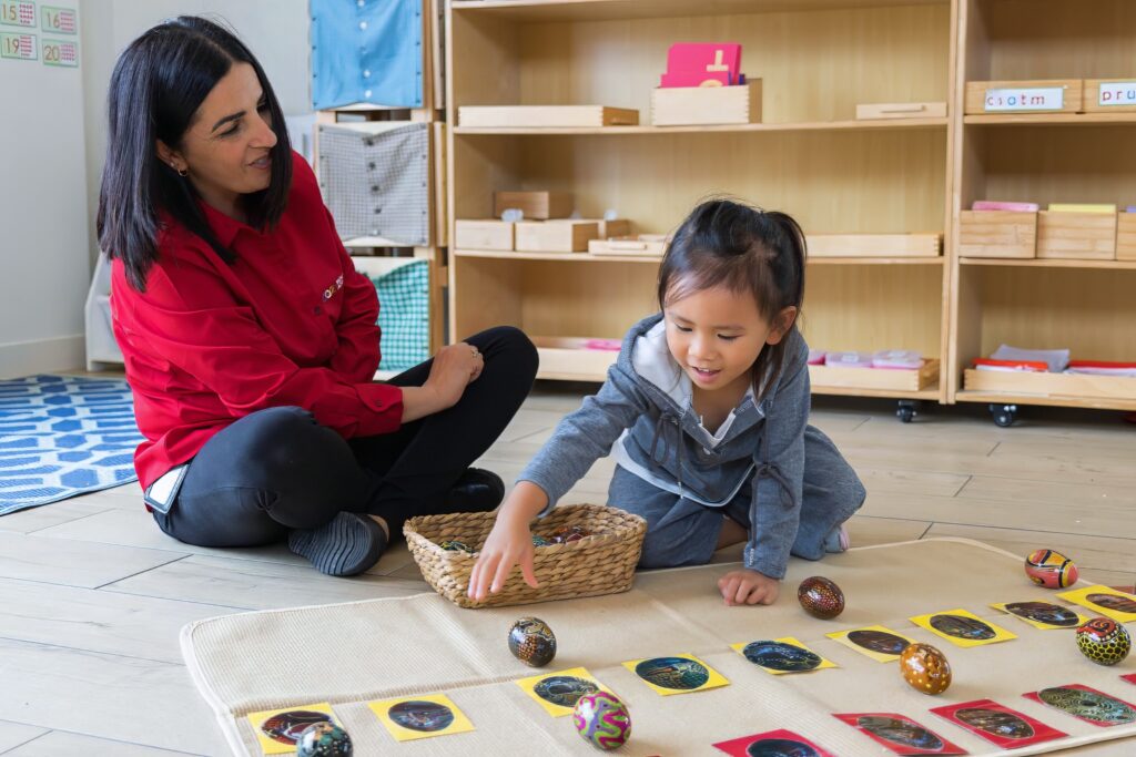 In a classroom, an educator sits cross-legged on the floor, observing a young girl who is matching a small object to its corresponding image on a work mat.