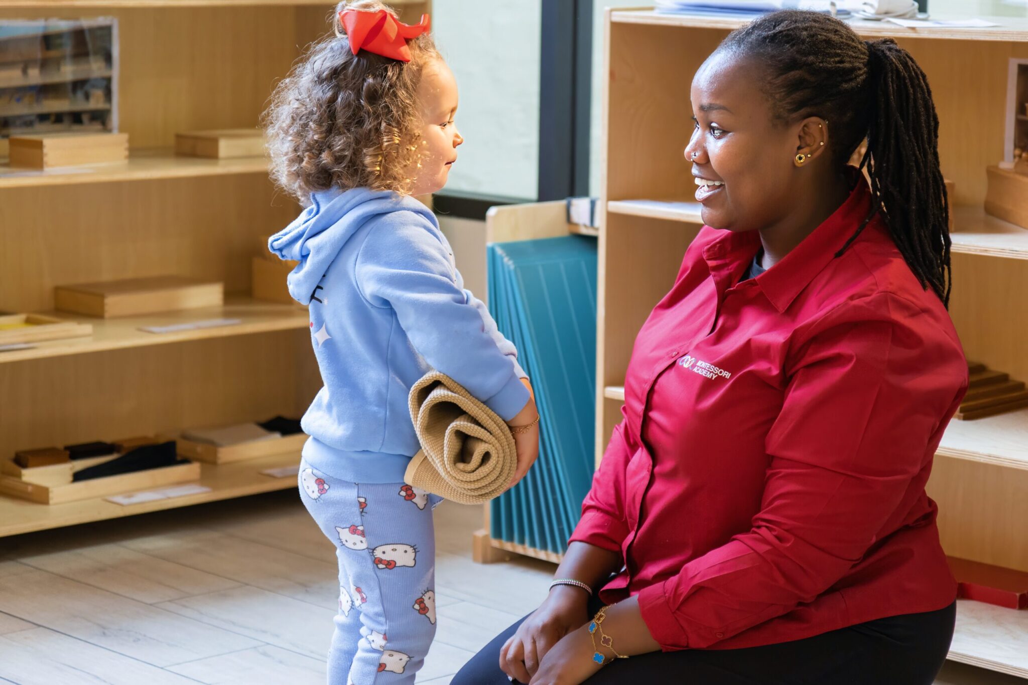 An educator kneels to meet a young child at eye level. They are both smiling warmly at each other, showing a moment of trust and connection.